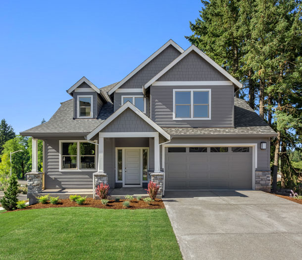 A light gray craftsman-style home with a green lawn and tall pine trees
