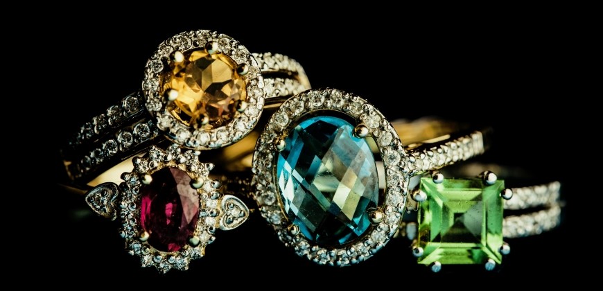 A red or pink sapphire ring, yellow sapphire ring, blue sapphire ring, and green sapphire ring stacked on a black background