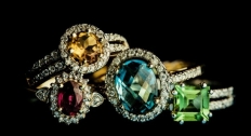 A red or pink sapphire ring, yellow sapphire ring, blue sapphire ring, and green sapphire ring stacked on a black background